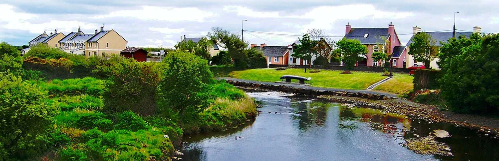 Main Street (N67) in Doonbeg, County Clare, Ireland, with a stream flowing past houses toward the Atlantic.